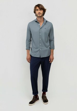 Hombre con cabello de longitud media que lleva una camisa azul claro abotonada, pantalones azul marino remangados en los tobillos, calcetines rojos y zapatillas marrones, posa sobre un fondo liso.