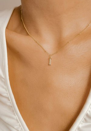 Halskette in Goldfarbe mit einem vertikalen Anhänger, der mit kleinen klaren Edelsteinen verziert ist, auf glatter Haut über einem weißen Kleidungsstück liegend.