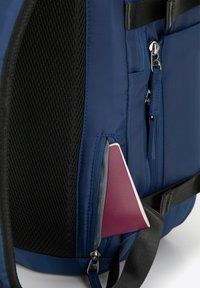 Tmavě modrý batoh s texturovanou síťovinou, kapsou na zip a viditelným vínovým chytrým telefonem uvnitř boční kapsy.