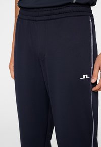 Marina atletiska byxor med dragsko i midjan, sidofickor och vitt logo. Har kontrasterande vita sömmar längs sidorna.