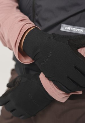 Person wearing black Ortovox gloves and a pink long sleeve shirt adjusting one glove on their left hand.