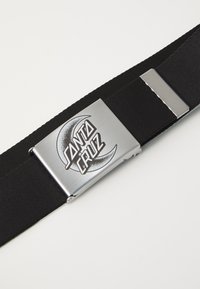 Melnas auduma jostas ar metalizētu sudraba sprādzi, kurā ir melns "SANTA CRUZ" logo un noapaļots akcenta dizains.