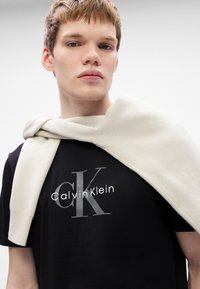 Melns T-krekls ar sudraba "Calvin Klein" logotipu. Krēmkrāsas adījums, kas karājas pār pleciem. Gluda tekstūra, minimālisma dizains.