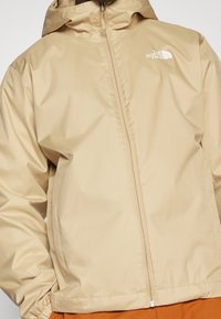 Beige zip-up jacket with hood, elastic cuffs, and white The North Face logo on the upper left chest.