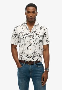 Camisa de manga corta en blanco con patrón de bambú negro, botonadura frontal, dos bolsillos en el pecho y botones beige. Llevada con jeans azules y un cinturón marrón.