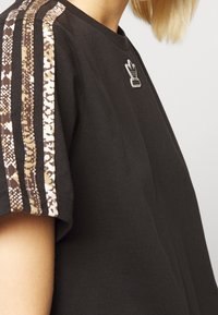 Černé bavlněné tričko s krátkým rukávem, hnědými vzorovanými pruhy na ramenech a kovovým logem Adidas na hrudi.