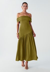 Schulterfreies grünes Kleid aus strukturiertem Stoff, tailliert mit ausgestelltem Rock und Falten, knöchellang. Getragen mit Riemchensandalen.