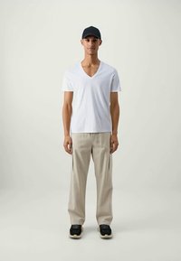 Jongeman met een zwarte pet, een witte V-hals T-shirt, beige wijde broek en zwarte sneakers, staand tegen een effen achtergrond.