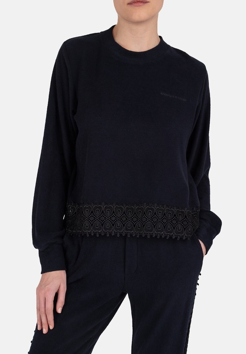Personne portant un haut noir à manches longues avec un bord en dentelle à l'ourlet et un pantalon noir assorti, les mains détendues le long du corps.