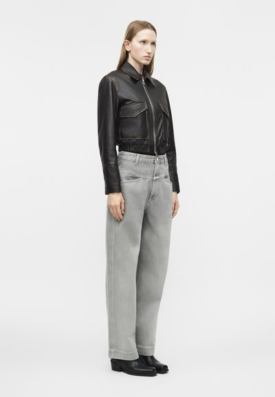 Woman standing wearing a fitted black leather jacket, wide-leg light gray jeans, and black heeled ankle boots against a plain background.