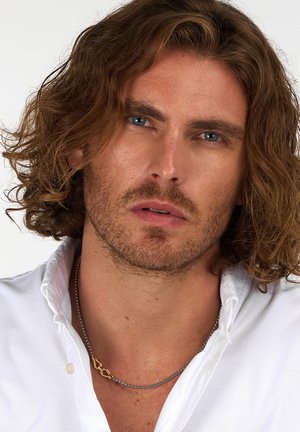 Modello maschile con capelli castani ondulati di lunghezza media, indossa una camicia bianca con bottoni e una collana di metallo argento con accenti dorati.