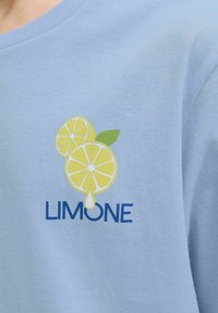 Šviesiai mėlyna medvilninė marškinėliai su grafika, vaizduojančia dvi geltonas citrinos riekeles ir žalią lapą, kartu su tekstu "LIMONE" mėlyna spalva.