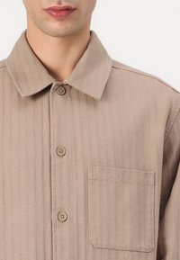 Camisa de algodón marrón con textura de canalé vertical, botón frontal y un bolsillo en el pecho. Presenta un cuello clásico y cuatro botones.