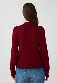 Maglione a maglia bordeaux con maniche lunghe, polsini a coste e scollatura rotonda. Bottoni a accento dorato ben visibili sui polsini delle maniche.