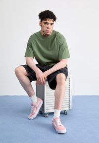 Grön bomulls-t-shirt, svarta denim-shorts, rosa mockasneakers med vita accenter, sittande på en metallvagn mot en ljus bakgrund.