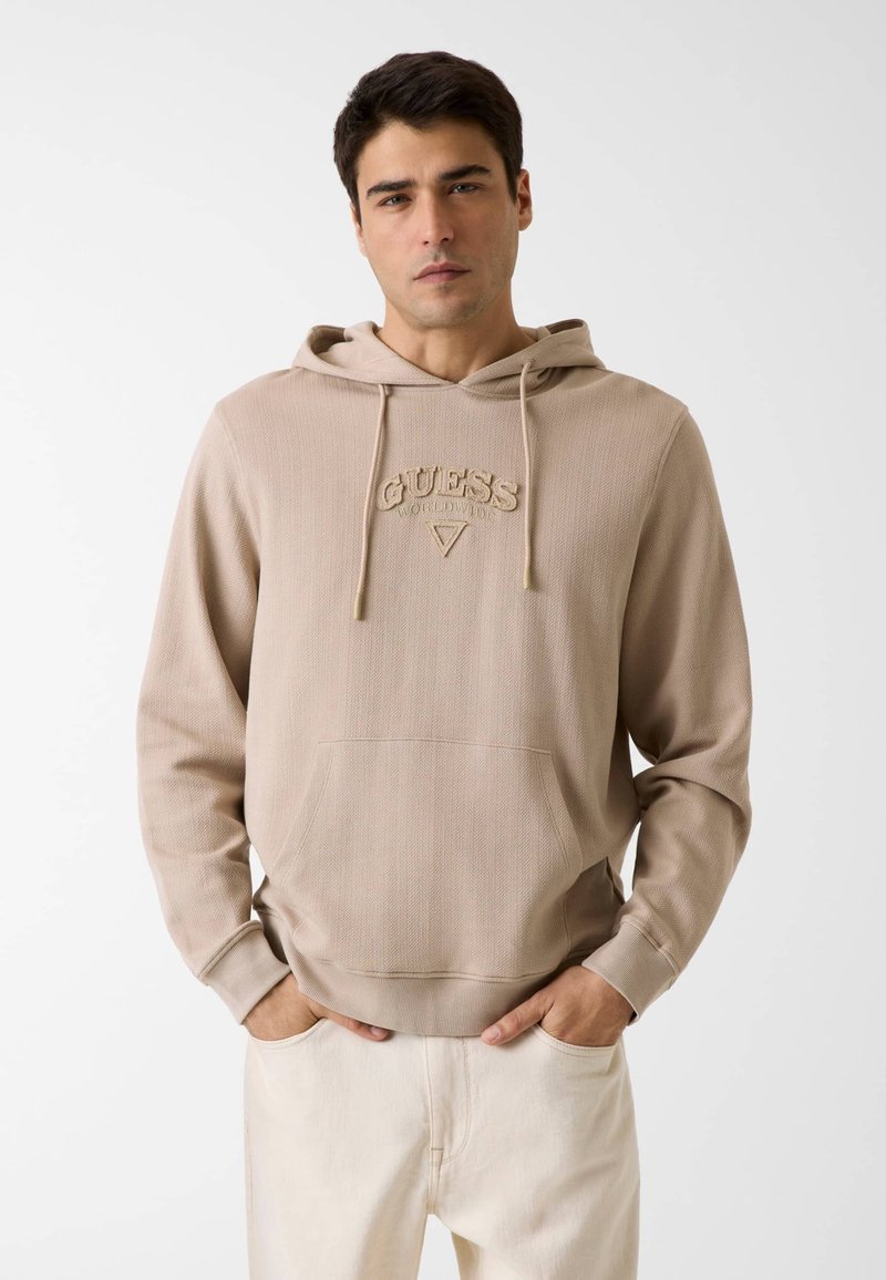 Jeune homme portant un sweat à capuche beige Guess avec poche avant et cordons, debout les mains dans les poches de son pantalon clair, sur un fond uni.