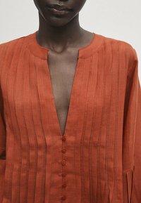 Rustfarvet linned blouse med lodrette striber, en dyb V-halsudskæring og syv matchende knapper ned ad fronten. Lange ærmer.