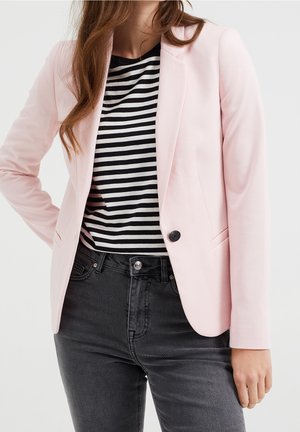 Femme portant un blazer rose clair sur une chemise rayée noir et blanc avec un jean taille haute noir, debout avec une main sur la hanche.