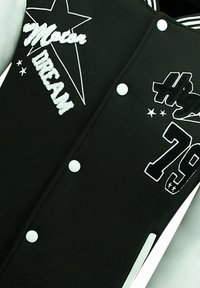 Zwarte varsityjack met witte geborduurde letters en accenten. Opmerkelijke details zijn een sterontwerp en drukknoopsluitingen. Rubberachtige patches zijn aanwezig.