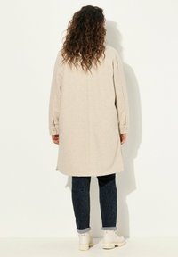 Béžový kabát z vlněné směsi s dlouhými rukávy, zadními rozparky a zapínacími manžetami, který je kombinován s tmavými vzorovanými džíny a světlými kotníkovými ботами.