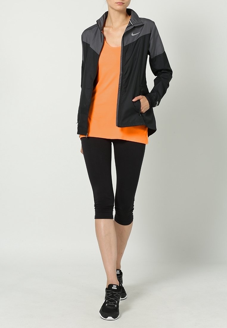 Giacca sportiva nera e grigia sopra una canotta arancione, abbinata a leggings capri neri e scarpe da corsa nere. Materiale liscio e traspirante.
