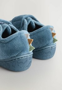 Des pantoufles en peluche bleues avec une texture douce, une forme arrondie et des accents en feutre triangulaires de couleur verte, marron, beige et grise à l'arrière.