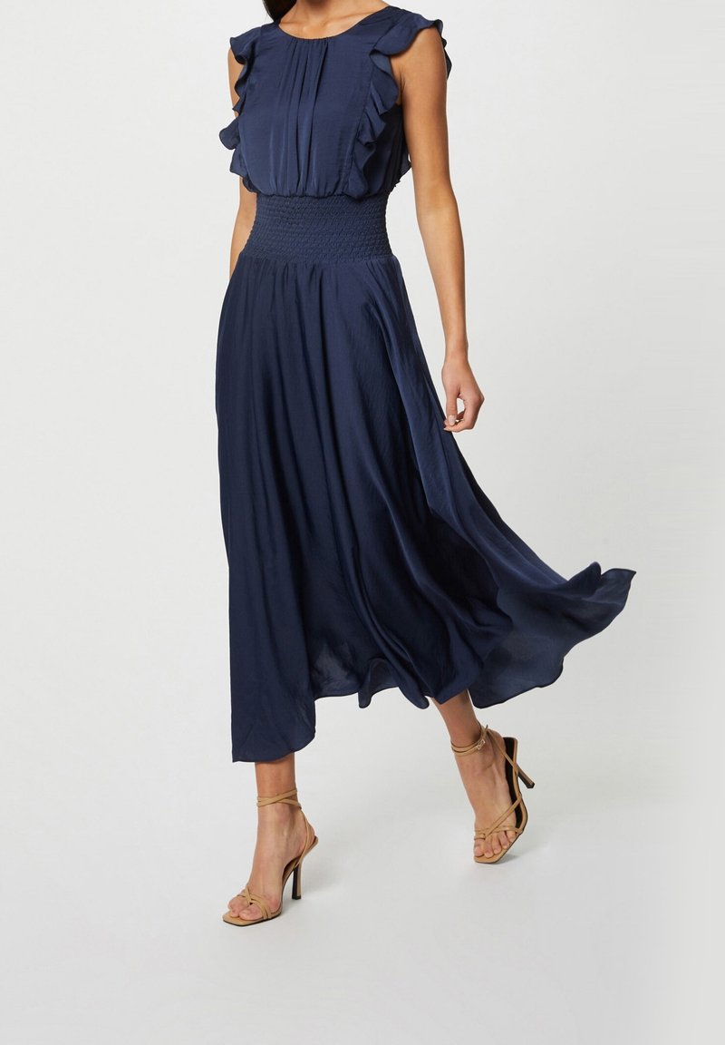 Kobieta w zwiewnej granatowej sukience midi z falbaniastymi rękawami i marszczoną talią, nosząca beżowe sandały na wysokim obcasie z paskami, idzie do przodu.