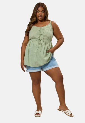 Femme portant un haut sans manches en vichy vert avec des détails à nouer, un short en denim bleu clair retroussé, et des sandales blanches à enfiler, debout avec une main dans la poche.