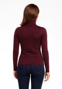 Maglione a maniche lunghe color bordeaux con collo alto, realizzato in tessuto elasticizzato, design aderente e texture liscia. Abbinato a jeans scuri.