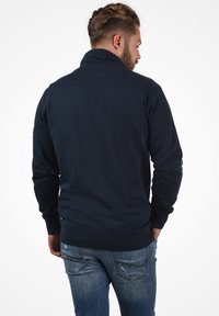 Sudadera azul marino con cuello alto, puños y dobladillo acanalados, textura suave. Llevada con jeans de mezclilla claros y desgastados. Vista trasera.