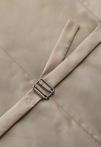 Tissu beige au toucher lisse, avec une boucle en métal et une sangle partiellement repliée. Les détails de couture sont visibles.
