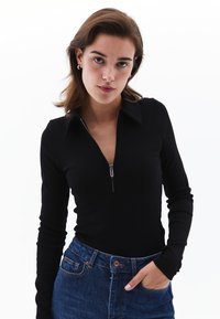 Schwarzer Reißverschluss-Bodysuit mit Kragen, langen Ärmeln, gerippter Textur und Slim-Fit, kombiniert mit blauen High-Waist-Jeans.