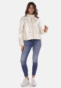 Giacca beige leggera e lucida con colletto alto, zip frontale e cordino. Abbinata a jeans blu e sneakers bianche.