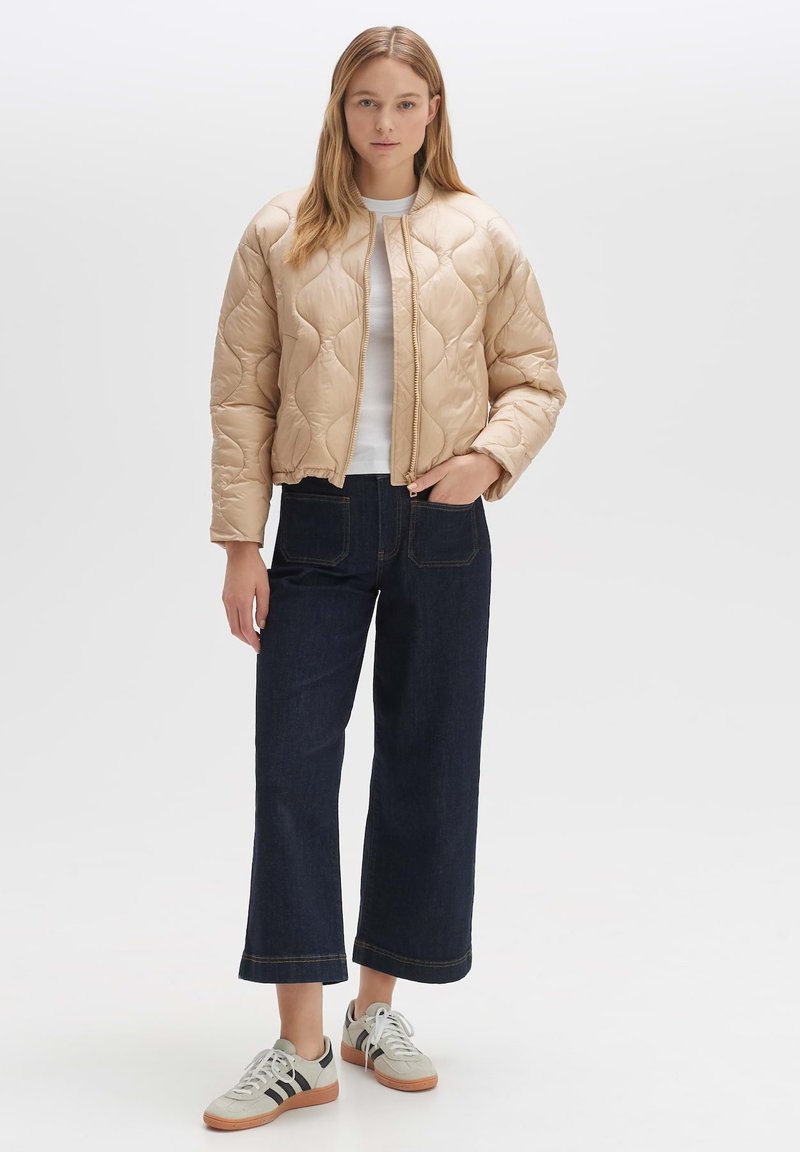 Beige Steppjacke mit einem gesteppten Muster, geripptem Kragen und Reißverschluss. Kombiniert mit dunklen, hochtaillierten Jeans und grauen Sneakers.