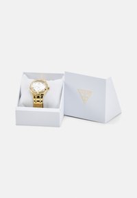 Montre en or avec un cadran rond, bracelet métallique et accents de pierres précieuses, placée sur un intérieur rembourré blanc dans une boîte grise clair avec un logo.