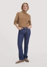 Beiger Rollkragenpullover, blaue Jeans und braune Ballerinas. Der Pullover hat lange Ärmel und gerippte Bündchen; die Jeans haben eine klassische gerade Passform.