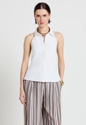 Frau trägt weiße ärmellose Bluse mit Kragen, gestreifte weit geschnittene Hose, goldenes Armreif und rundes Anhängerhalskette vor schlichtem Hintergrund.