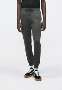 Man in donkere grijze joggingbroek met een trekkoord in de taille en logo op de dij, zwarte sneakers met witte veters en witte sokken, staande op een witte achtergrond.