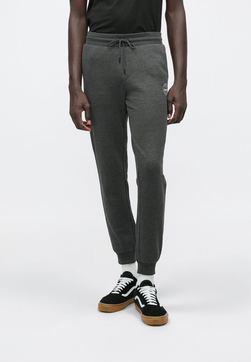 Man in donkere grijze joggingbroek met een trekkoord in de taille en logo op de dij, zwarte sneakers met witte veters en witte sokken, staande op een witte achtergrond.