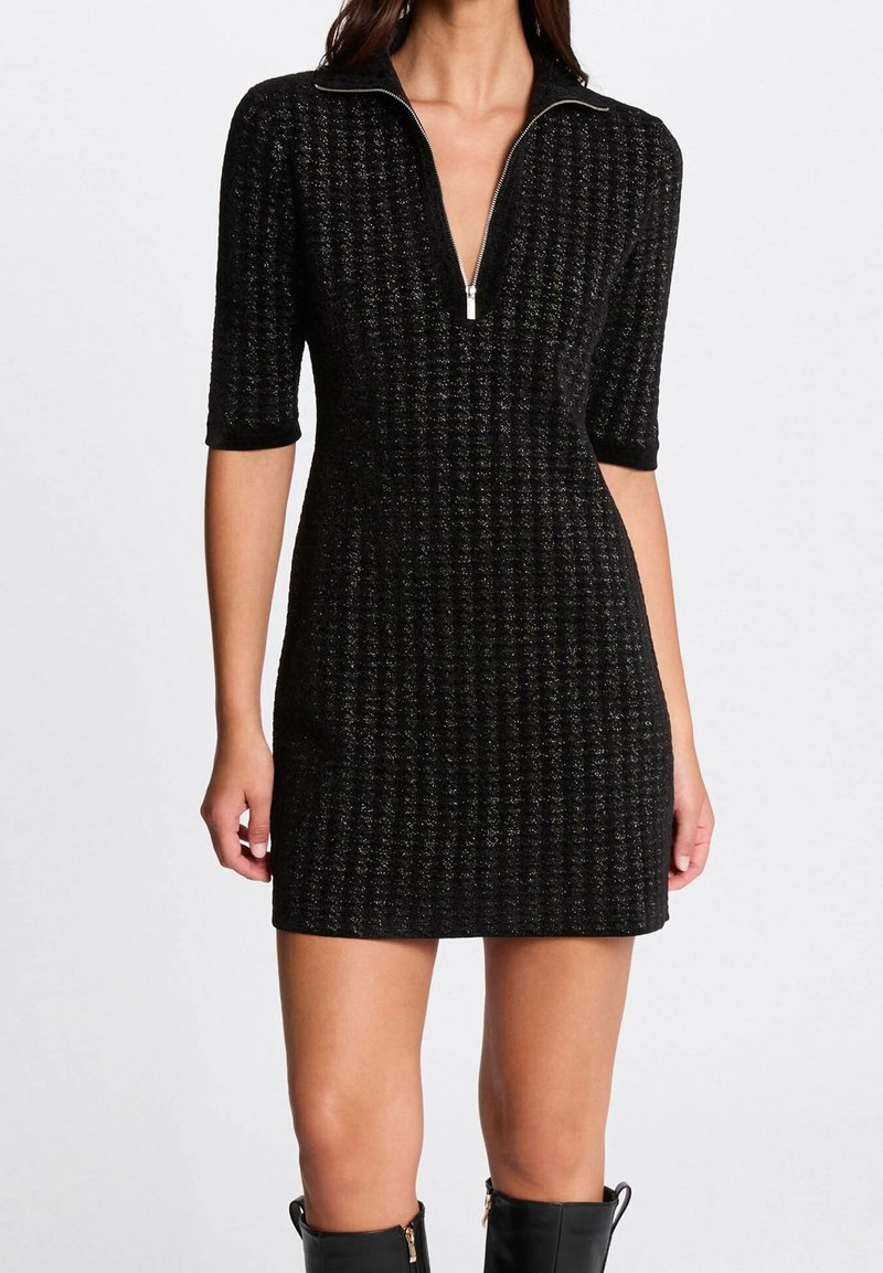 Femme portant une mini-robe en maille texturée noire avec des manches jusqu'au coude, une fermeture éclair argentée au col et des bottes noires montantes jusqu'aux genoux.