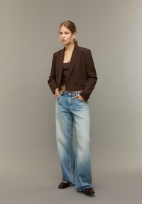 Bruine cropped blazer over een bruin topje, gecombineerd met lichtblauwe wijde jeans. Het model staat zelfverzekerd met haar handen in de zakken, op een neutrale achtergrond.