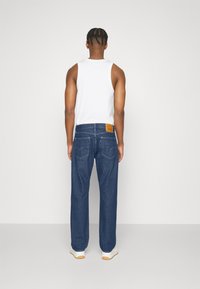 Jeans de mezclilla en un lavado azul oscuro, de corte holgado, con un parche de cuero marrón claro en la parte trasera, combinados con una camiseta sin mangas blanca y zapatillas blancas.
