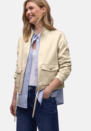 Femme souriante portant une veste zippée beige clair sur une chemise rayée bleue et blanche et un jean bleu foncé, avec une main dans la poche.