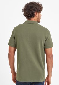 Man with curly hair wearing an olive green short-sleeve polo shirt and blue jeans, facing away on a white background.