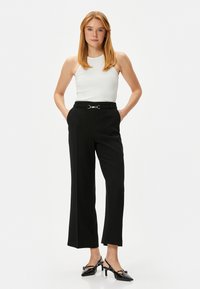 Zwarte wijde broek met een riem met een zilveren ringdetail, gedragen met een strakke witte mouwloze top en zwarte hoge hakken.