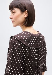 Jeune femme aux cheveux courts et foncés, portant une robe marron à pois blancs avec un col à volants, tournée vers la gauche sur un fond uni.