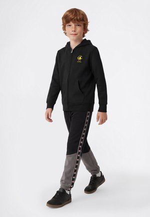 Roodharig jongetje dat loopt, draagt een zwart zip-up hoodie met een geel logo en zwarte-grijze trainingsbroek met zijlogo strepen en zwarte sneakers.