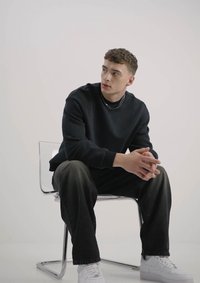 Schwarzer Pullover mit Rundhalsausschnitt, dunkle Hose und weiße Sneakers. Sitzt auf einem transparenten Stuhl, mit einer sichtbaren Kettenhalskette.