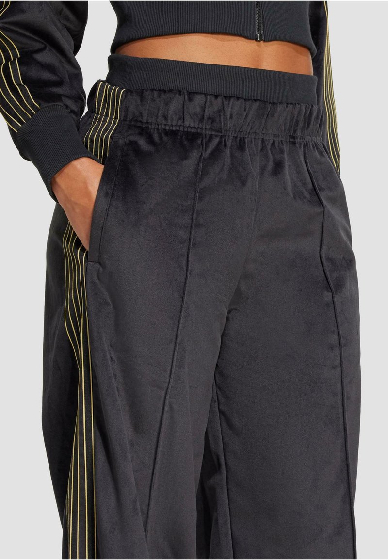 Pantaloni jogger neri in satin con strisce verticali gialle, vita elastica e tasche laterali. Tessuto liscio, vestibilità adattata e gambe affusolate.