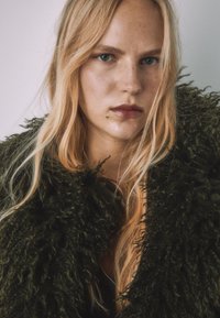 Zeleni, puhasti kožuh z zgrbljeno teksturo z visokim ovratnikom in zadrgo. Dolgi, ravni lasje delno prekrivajo obraz.